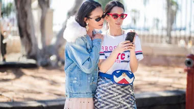 Two stylish women wearing sunglasses, looking at a smartphone outdoors.