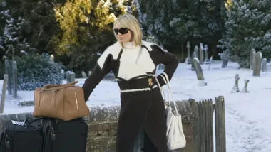 Blonde woman in a stylish coat and sunglasses stands beside luggage in a snowy cemetery.