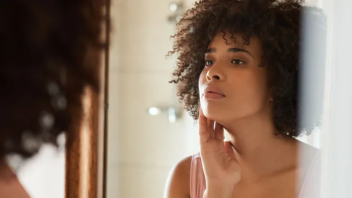 Woman with curly hair touches her chin while looking into a mirror.