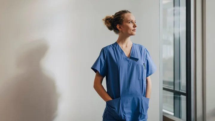 A healthcare worker in blue scrubs stands with hands in pockets, looking out a window.