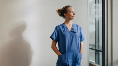 A healthcare worker in blue scrubs stands with hands in pockets, looking out a window.