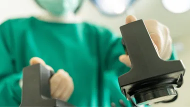 Surgeon in green scrubs holding defibrillator paddles, with a blurred background of operating room lights.