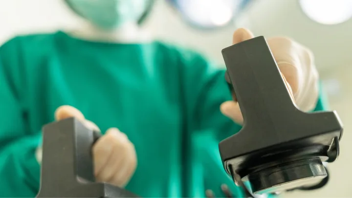 Surgeon in green scrubs holding defibrillator paddles, with a blurred background of operating room lights.