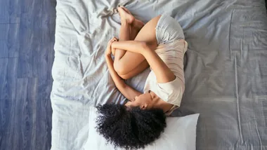 A person lying on a bed in a fetal position, wearing a light shirt and shorts, with curly hair spread on the pillow.