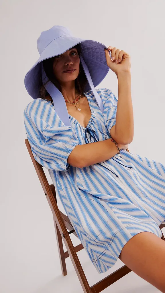 A woman wearing a light blue pastel hat modelling for brand Free People. The model sits on a fold out chair wearing a blue striped mini dress.