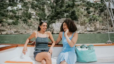 Two women laughing and enjoying a sunny day on a boat with a scenic rocky hillside in the background.