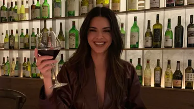 A person smiling while holding a glass of red wine in a room with shelves of wine bottles in the background.