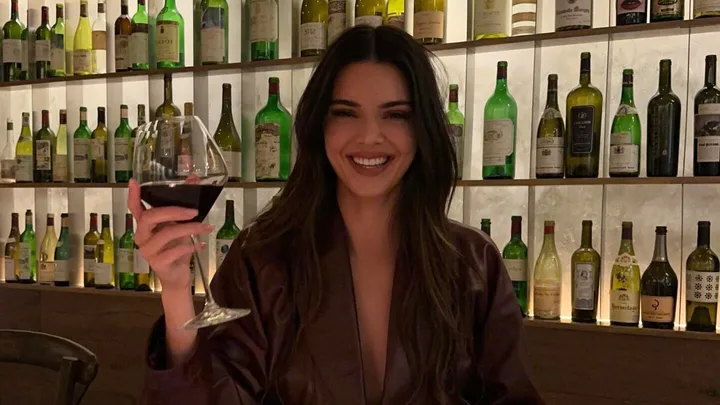 A person smiling while holding a glass of red wine in a room with shelves of wine bottles in the background.