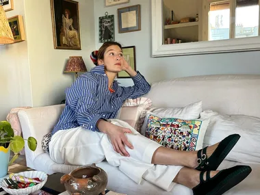 A person in a striped shirt and white pants sits on a sofa, surrounded by framed art on the wall and a coffee table with magazines.