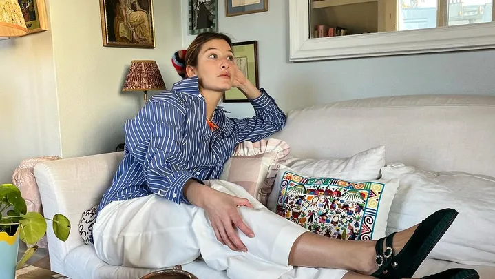 A person in a striped shirt and white pants sits on a sofa, surrounded by framed art on the wall and a coffee table with magazines.