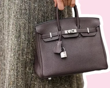Brown leather handbag being held by a person wearing a textured skirt, with a pink background.