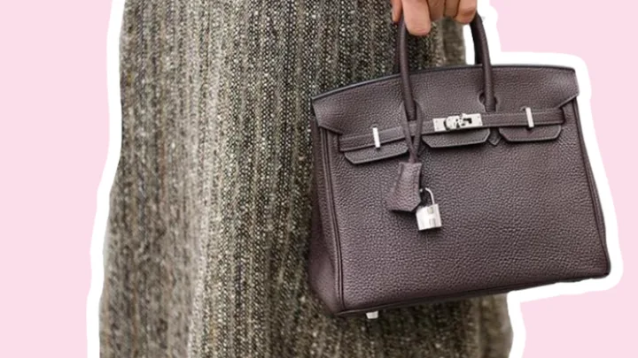 Brown leather handbag being held by a person wearing a textured skirt, with a pink background.