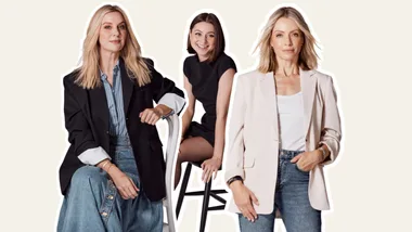 Three women posing against a plain background, each wearing blazers and jeans, with one sitting on a high stool.