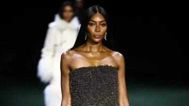 Model walks runway in a strapless textured black dress, with long hair and earrings, another model blurred in the background.