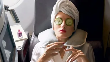 Woman in a plane seat, with towel on head, cucumber slices on eyes, and painting nails; red nail polish bottle on table.