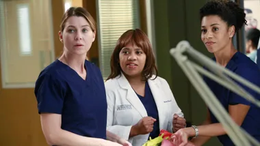 Three women in medical scrubs and a lab coat standing in a hospital setting, focused on something unseen.