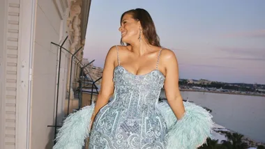 Woman in a blue sequined dress with feathered sleeves smiling on a balcony overlooking a coastal city at sunset.