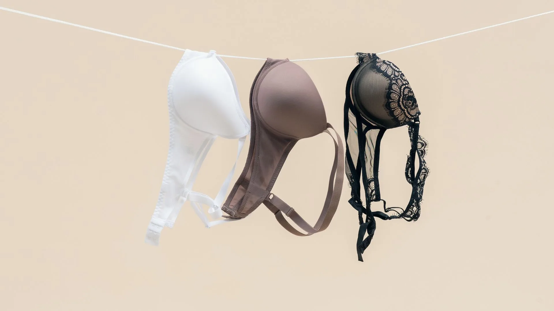 Three bras in white, brown, and black lace hang on a line against a soft beige background.