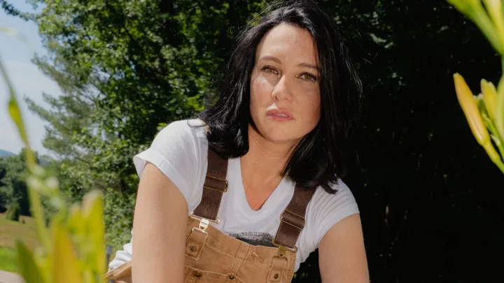 A person with black hair wearing a white t-shirt and brown overalls, outdoors with greenery in the background.