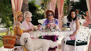 Four women in vibrant 1960s attire sit at an outdoor table under striped canopies with a white poodle nearby.