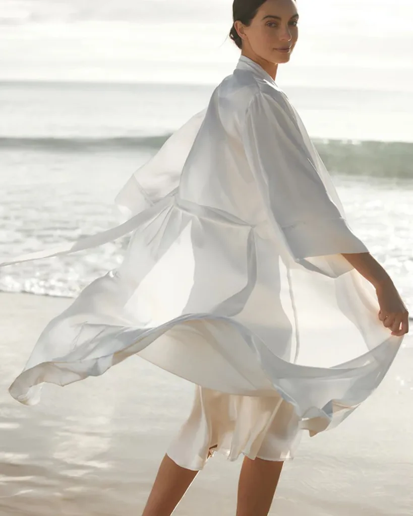A woman standing on the shoreline of a beach wearing a white silk nightie and robe.