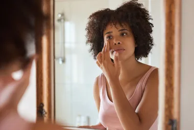 Young African woman putting moisturizer on her face while getting ready in her bathroom in the morning