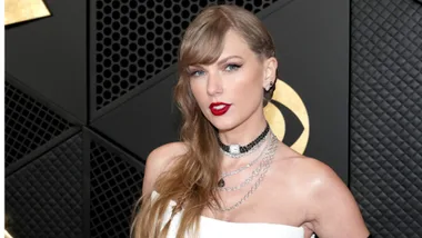 A person with long hair and red lipstick, wearing a white outfit and layered necklaces at an event.