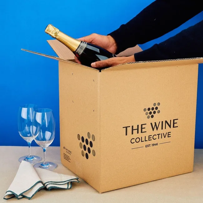 Opening a wine bottle in a "The Wine Collective" box with two glasses and napkins nearby.