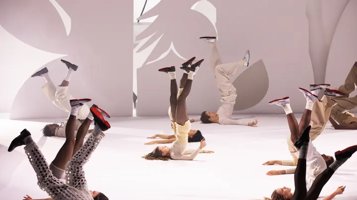 Dancers lying on stage with legs raised, wearing red-soled shoes.