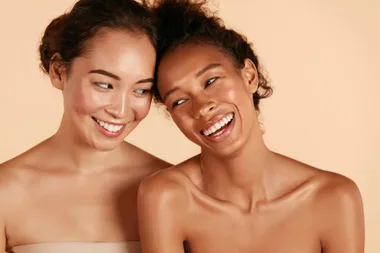 Two women with glowing skin laughing together against a beige background.