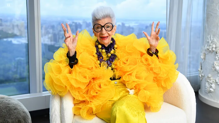 Elderly person with gray hair, large glasses, wearing a vibrant yellow outfit, sitting indoors with a city view behind.