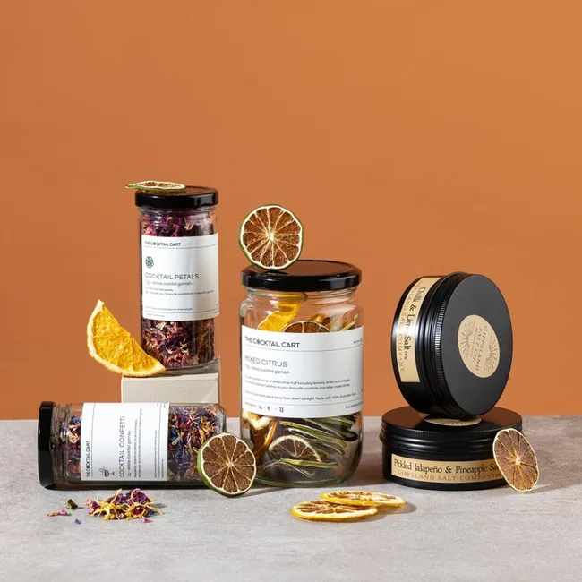 Assorted jars of The Cocktail Cart dried garnishes, including citrus slices and petals, arranged on a surface with an orange background.