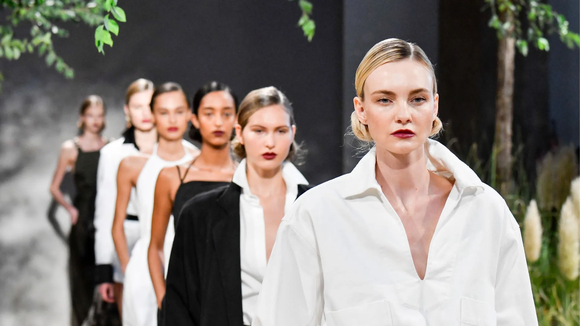 Models walking down runway in fashion show, diverse outfits, lush background.