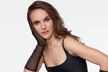 A woman in a black dress and sheer black glove poses with her hand on her face against a plain background.