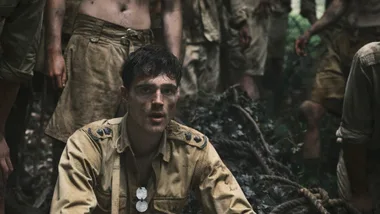 Young soldier in a worn uniform, with a dirt-smeared face, sits in a jungle setting, surrounded by others in similar attire.