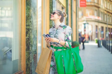 Woman in floral dress shopping outdoors, holding bags and a phone, looking up and smiling.