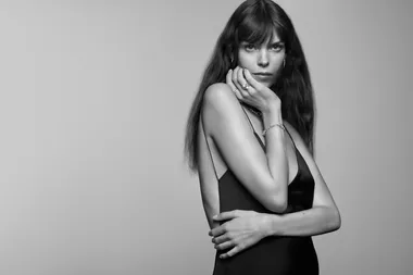 Black and white photo of a woman with long hair in a sleeveless dress, posing with hand on chin, looking at the camera.
