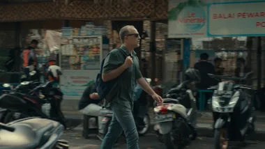 Man walking with a backpack on a bustling street with motorbikes, wearing sunglasses and green shirt.