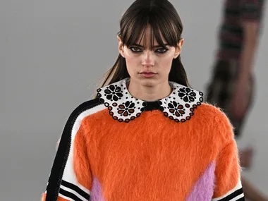 Model with long brown hair wearing an orange fuzzy sweater and black skirt on the runway.