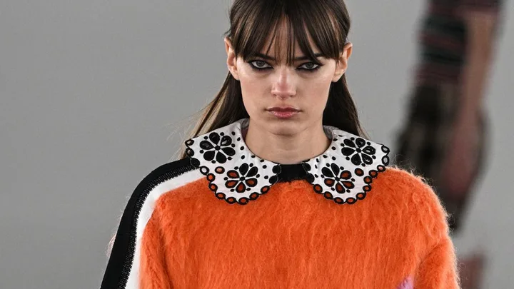 Model with long brown hair wearing an orange fuzzy sweater and black skirt on the runway.