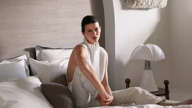 Woman in a white sleeveless top sitting on a bed next to a modern white lamp.
