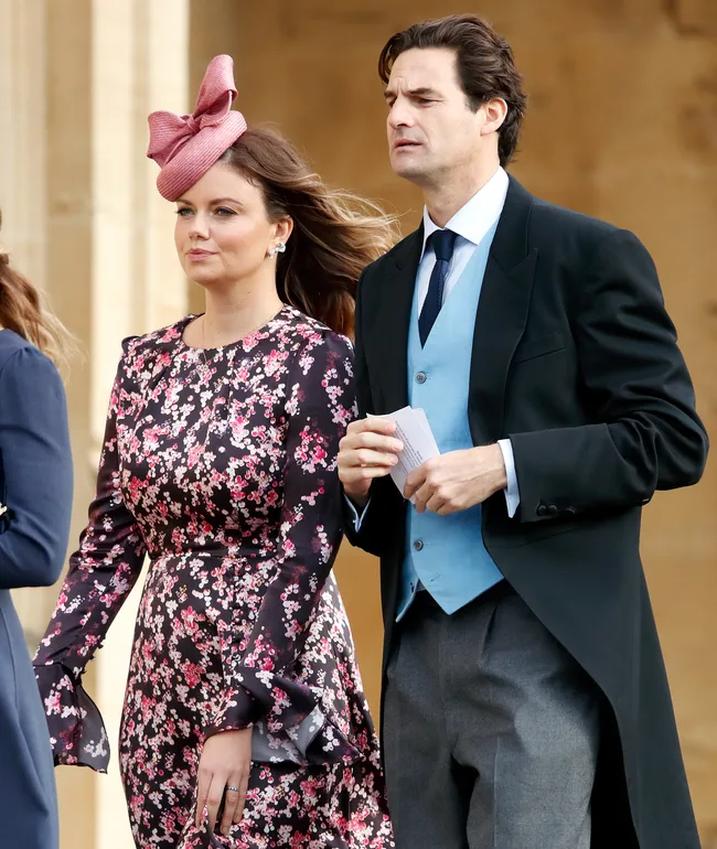 A woman in a floral dress with a pink fascinator and a man in a suit with a blue vest walking together outdoors.