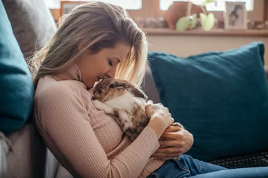 Woman hugging a rabbit while sitting on a couch, with cozy indoor decor.