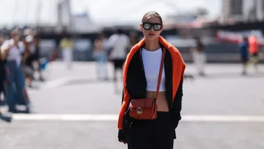 woman stands in street with orange jumper over her shoulders and a crossbody bag