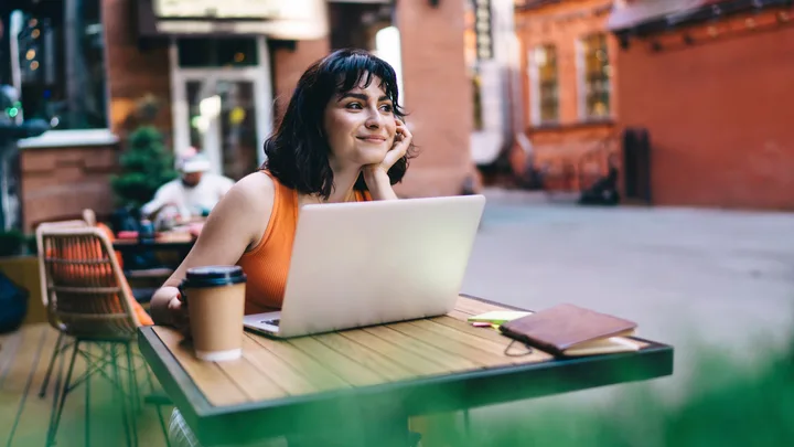 Woman in orange top sits at outdoor café table, smiling at laptop with coffee and notebook nearby.
