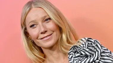 A person with long blonde hair smiles, wearing a striped outfit against a peach background.