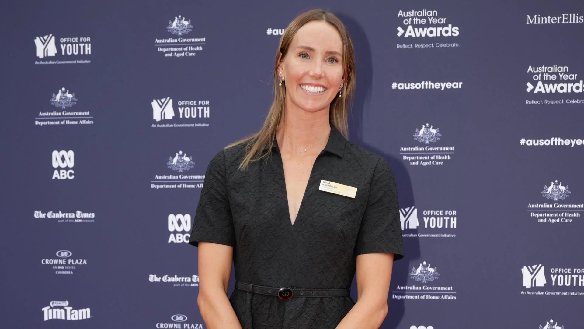 A woman in a black dress smiles in front of a backdrop with Australian of the Year Awards logos.