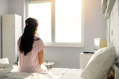 A woman in a hospital gown sits on a bed, facing a window with sunlight streaming in.