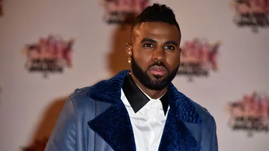 Man in a blue jacket with a beard stands against a blurred award show backdrop.