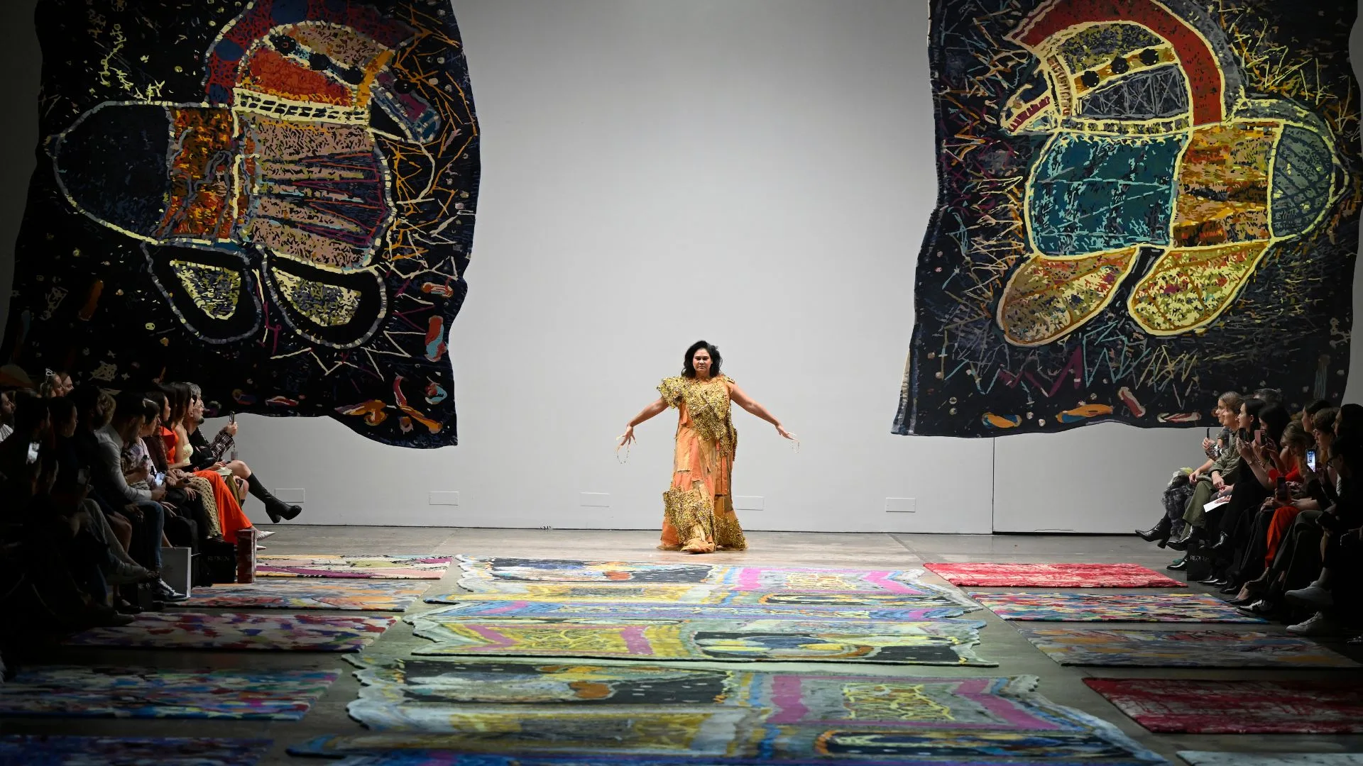 Model walks runway with vibrant abstract art; audience sits on either side.